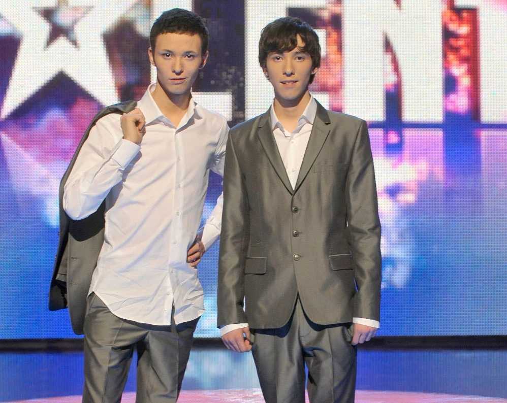 Bracia Legunowie w programie "Mam talent" w 2009 r. Dwóch młodych mężczyzn ubranych elegancko w garnitury pozuje na tle świetlistej, scenicznej planszy, jeden z nich trzyma marynarkę zarzuconą na ramię.