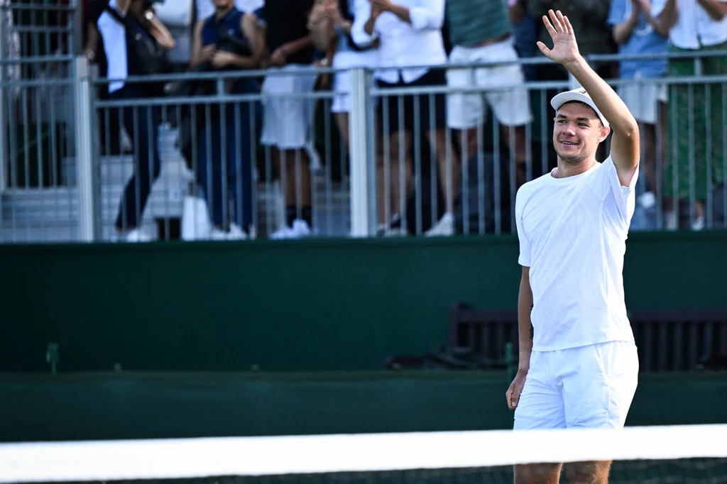Kamil Majchrzak niebawem zagra o ćwierćfinał Wimbledonu. Jeszcze w piątek poznał najbliższego rywala Tenisista w białym stroju stoi na korcie, uśmiecha się i macha ręką do publiczności, za nim widoczni są kibice stojący przy ogrodzeniu.