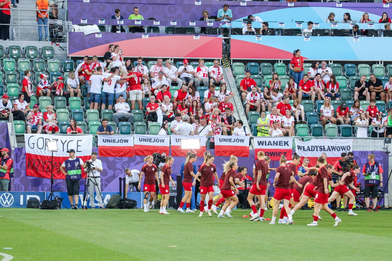 Reprezentacja Polski na rozgrzewce przed debiutem na Euro 2025 Piłkarki w czerwonych strojach zbierają się na murawie stadionu, wokół widoczne są trybuny z liczną grupą kibiców ubranych w biało-czerwone barwy, na ogrodzeniu wywieszone są polskie flagi i transparenty z nazwami miejscowości.