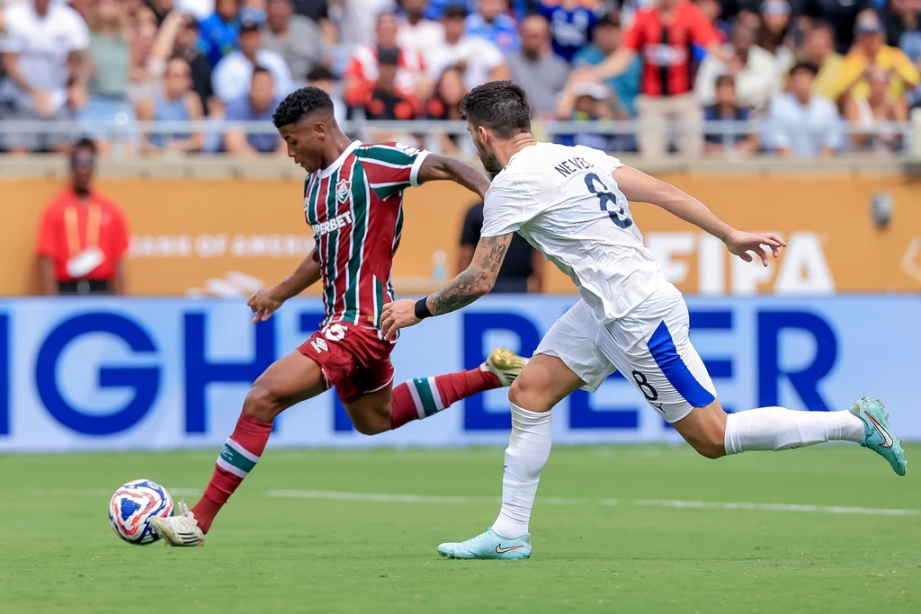 Fluminense - Al-Hilal Dwóch piłkarzy na boisku w trakcie dynamicznej akcji, zawodnik w pasiastym stroju wykonuje strzał lub podanie, zawodnik w białym stroju próbuje go powstrzymać, w tle trybuny z kibicami.