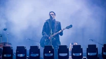 Muzyk grający na gitarze elektrycznej i śpiewający do mikrofonu na scenie, otoczony niebieskim światłem oraz mgłą, z widocznymi reflektorami w pierwszym planie.