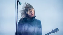 Gitarzysta z długimi rozwianymi włosami gra na gitarze elektrycznej podczas koncertu na scenie, dynamiczne światło oraz poruszające się włosy podkreślają energię występu