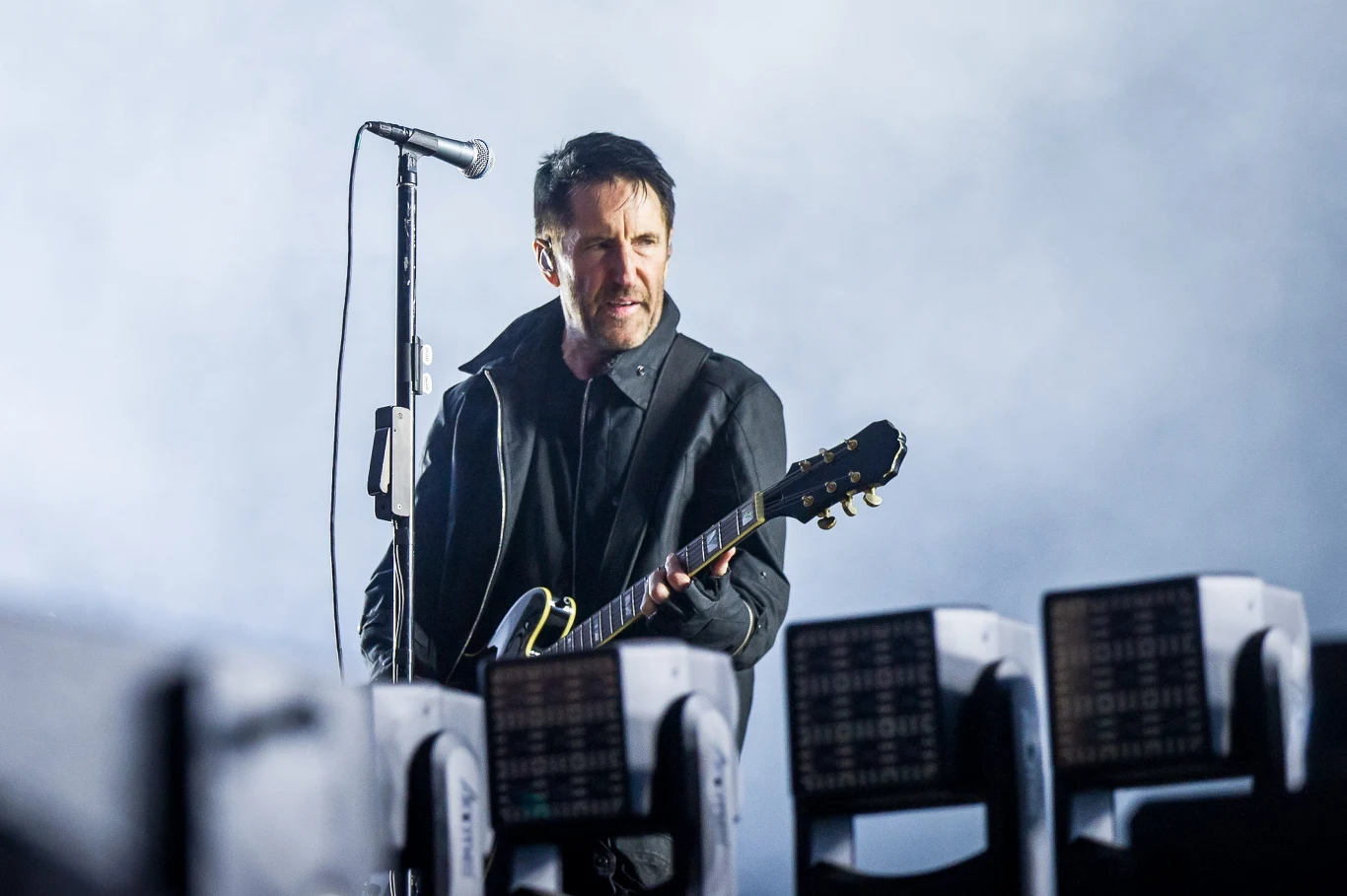 Nine Inch Nails na Open'er Festival 2025 Muzyk z gitarą elektryczną występuje na scenie, trzyma mikrofon i jest oświetlony jasnym światłem, w tle widoczny dym lub mgła sceniczna oraz reflektory.