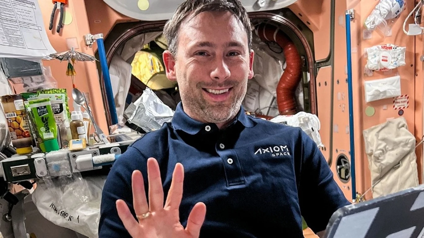 Spotkanie na żywo z polskim astronautą w Rzeszowie Mężczyzna w koszulce z logo 'Axiom Space' unosi rękę w geście powitania w otoczeniu wnętrza stacji kosmicznej, wokół niego widoczne są różne przyrządy oraz sprzęt do pracy w kosmosie.
