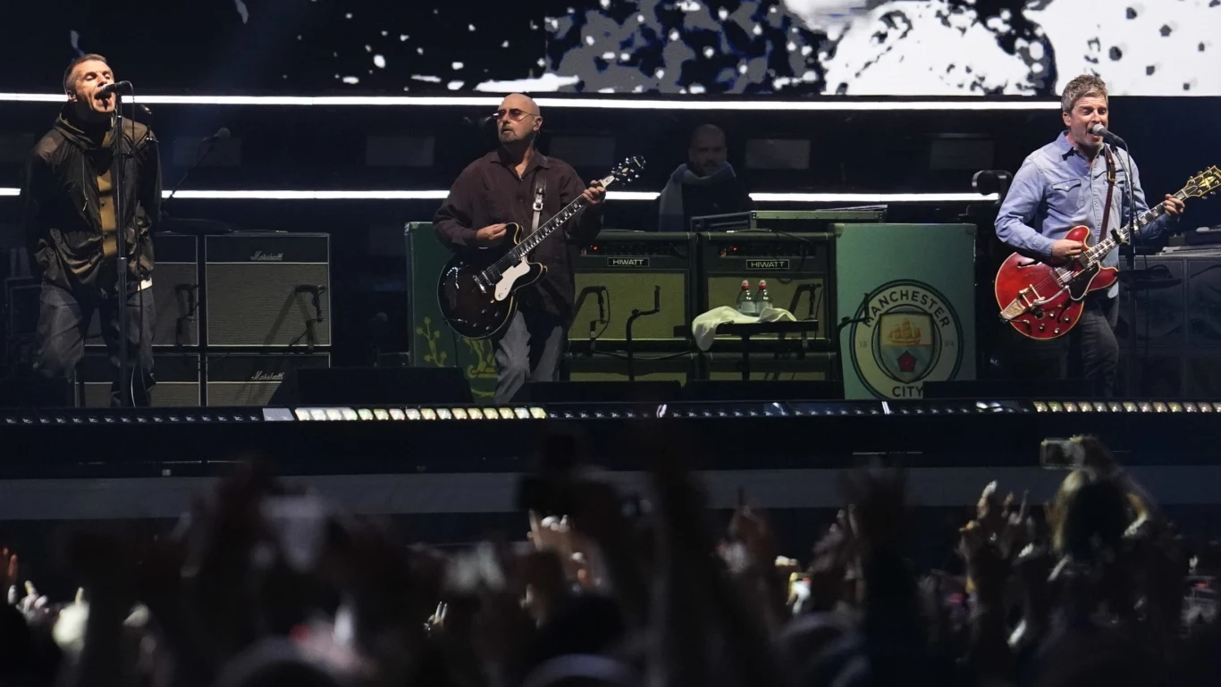 Oasis po latach wrócili na scenę Trzech muzyków wykonujących utwory na dużej scenie podczas koncertu, z tłumem uniesionych rąk na pierwszym planie oraz wizualizacjami graficznymi z tyłu sceny, na której znajduje się herb Manchester City.