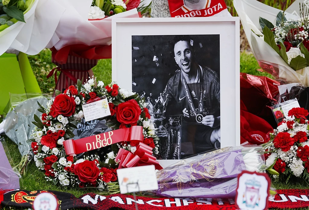 Kibice honorują zmarłego Diogo Jotę przed Anfield, 04.07.2025 r. Czarno-biała fotografia piłkarza otoczona kwiatami, wieńcami oraz szalikami klubowymi, tworzącymi miejsce pamięci. Główne elementy to uśmiechnięty sportowiec na zdjęciu i licznie poukładane czerwone róże.