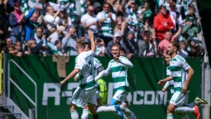 Czterech piłkarzy drużyny w biało-zielonych strojach świętuje zdobycie bramki podczas meczu piłki nożnej, w tle liczna, wiwatująca publiczność na trybunach.