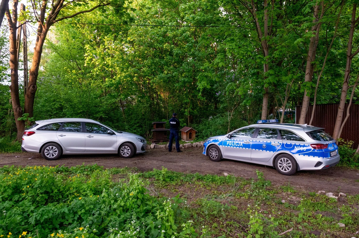 Policjant stoi przy dwóch samochodach, jeden z nich oznakowany jako radiowóz policyjny, drugi prywatny, zaparkowanych na leśnej drodze otoczonej gęstą zielenią. W tle widoczne są drewniane budy dla zwierząt.