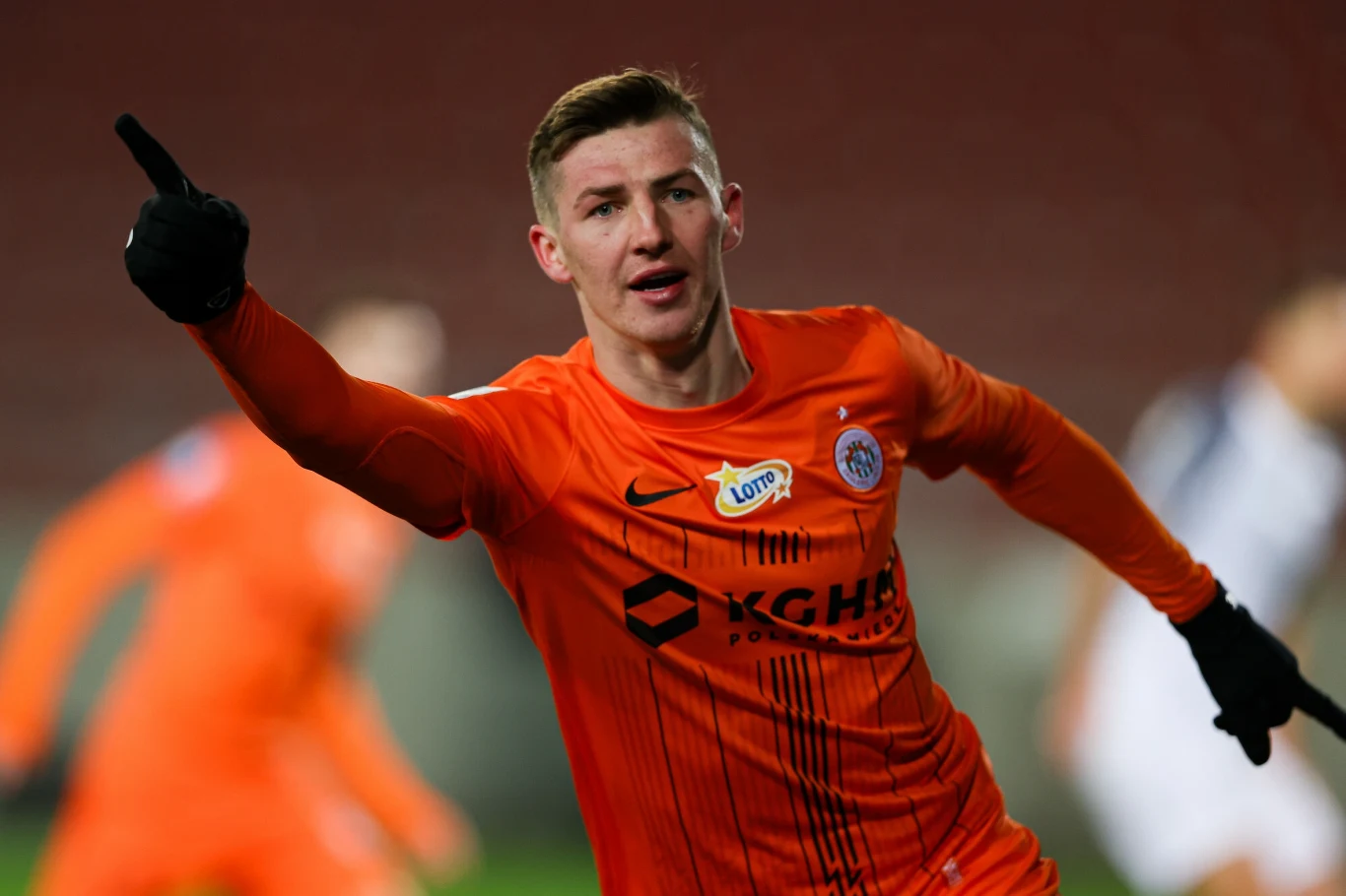 Dawid Kurminowski Piłkarz w pomarańczowym stroju z logo KGHM Zagłębie Lubin świętuje zdobycie bramki, unosi rękę z wyciągniętym palcem wskazującym, w tle nieostre postaci i stadion.