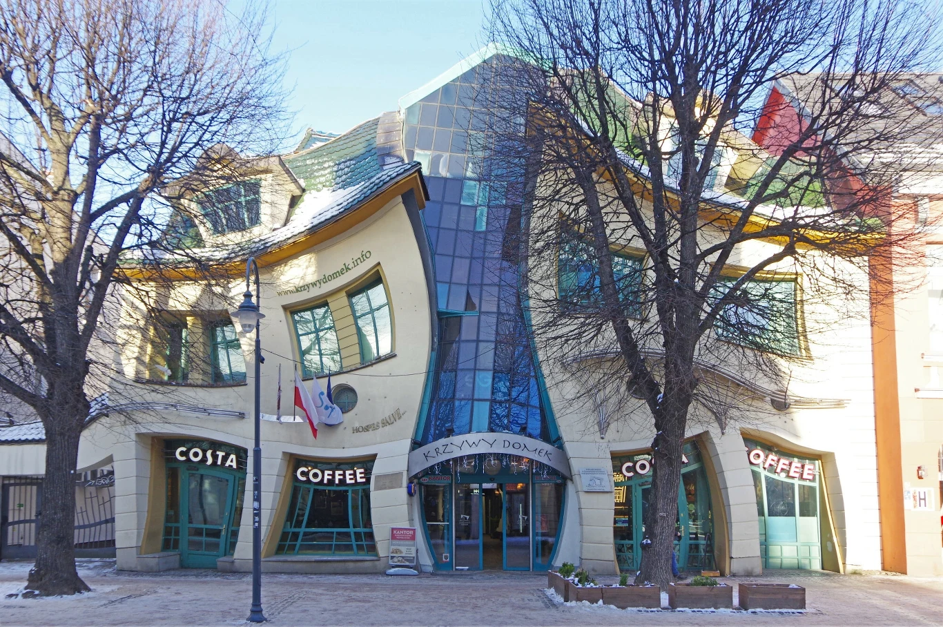 Budynek o charakterystycznej, falistej i zdeformowanej architekturze, znany jako Krzywy Domek, z dużymi przeszkleniami, tabliczką Costa Coffee oraz polską flagą, otoczony bezlistnymi drzewami.