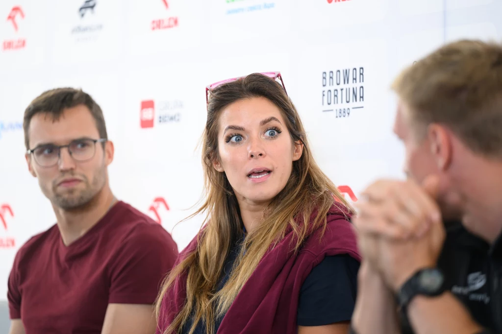 Maria Anddrejczyk Trzy osoby siedzące przy stole w rozmowie, kobieta na środku z wyraźnym, zaskoczonym wyrazem twarzy, mężczyzna po lewej patrzy w jej kierunku, a osoba po prawej zwrócona częściowo bokiem.