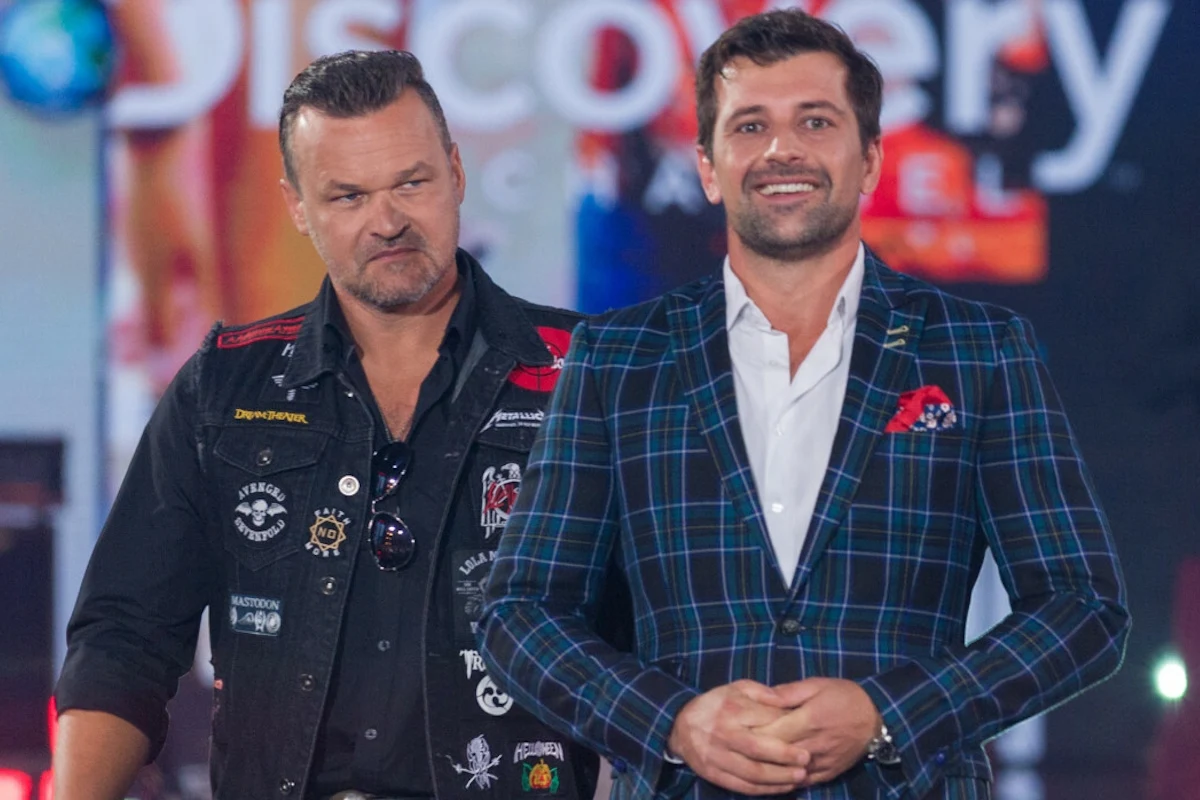 Patryk Mikiciuk i Adam Kornacki zaprzeczają stawianym im zarzutom Dwóch mężczyzn stoi razem w jasnym świetle, jeden ubrany w czarną kurtkę ozdobioną naszywkami, drugi w niebieską kraciastą marynarkę z poszetką, w tle widoczne kolorowe logo Discovery Channel.