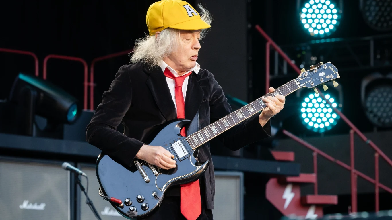 AC/DC zagrali na PGE Narodowym! Gitarzysta w czarnym garniturze, czerwonym krawacie i żółtej czapce gra na elektrycznej gitarze na scenie koncertowej, za nim widać wzmacniacze i sceniczne oświetlenie.