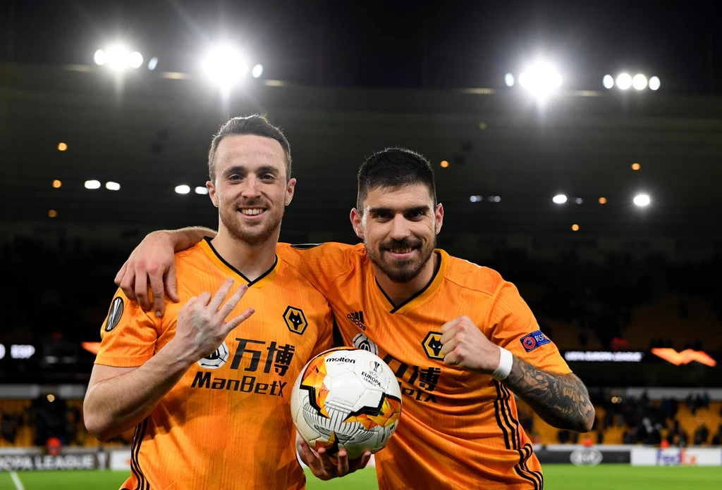 Diogo Jota i Ruben Neves Dwóch piłkarzy w pomarańczowych koszulkach drużyny Wolverhampton Wanderers stoi razem na stadionie, uśmiechając się i świętując zwycięstwo, jeden z nich trzyma piłkę meczową.