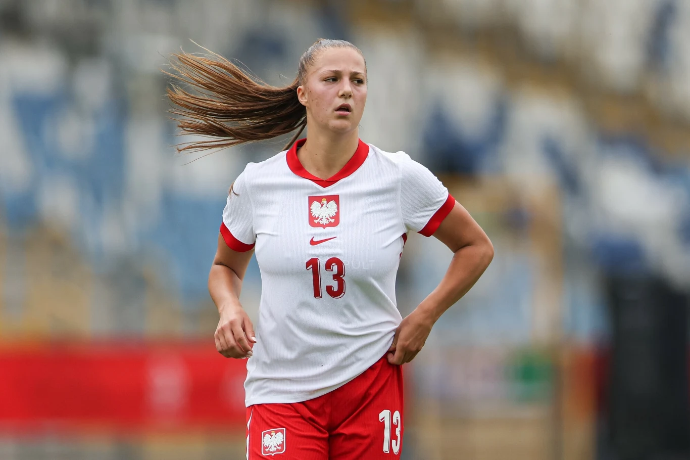 Piłkarka w stroju reprezentacji Polski z numerem 13 na koszulce biegnie po boisku sportowym podczas meczu, w tle widoczne rozmyte trybuny stadionu.