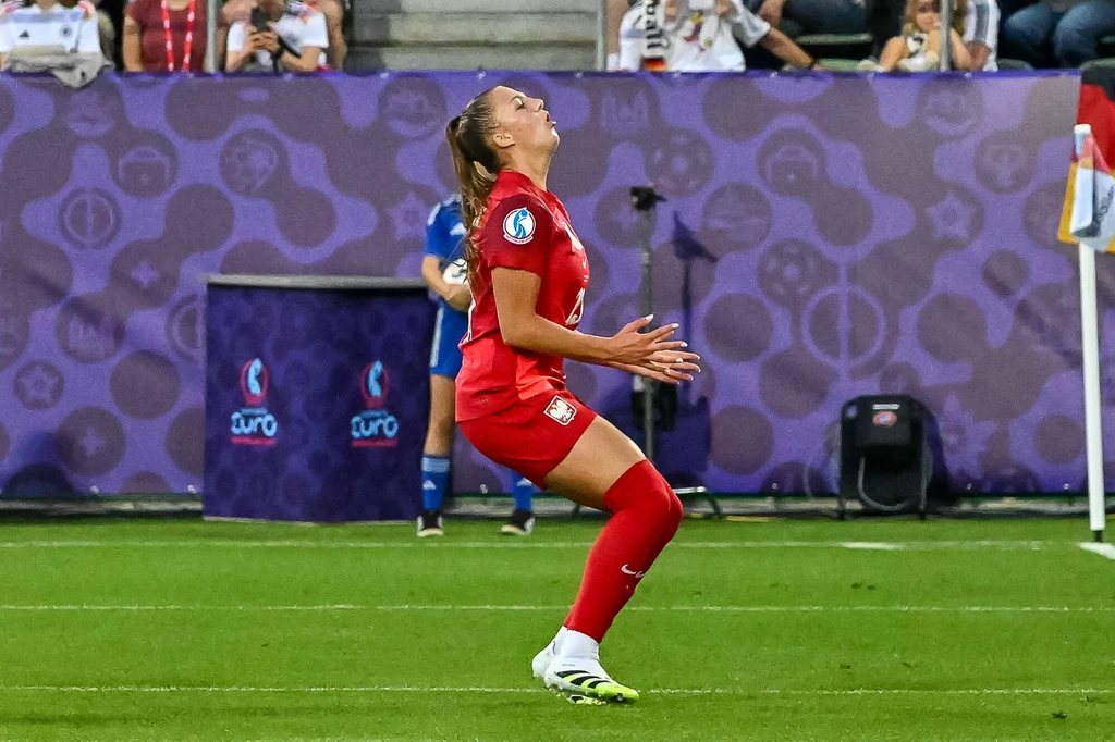 Emilia Szymczak Piłkarka ubrana w czerwony strój sportowy znajduje się na boisku piłkarskim podczas meczu, wykonując dynamiczny ruch z głową odchyloną do tyłu, wokół widoczna jest murawa i fragment trybun z kibicami.