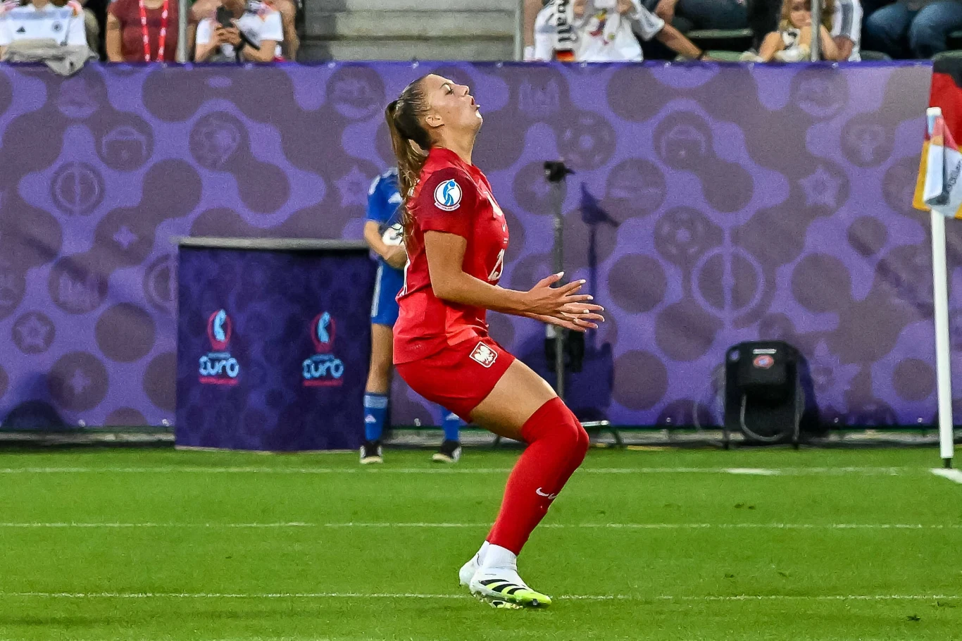 Piłkarka ubrana w czerwony strój sportowy znajduje się na boisku piłkarskim podczas meczu, wykonując dynamiczny ruch z głową odchyloną do tyłu, wokół widoczna jest murawa i fragment trybun z kibicami.