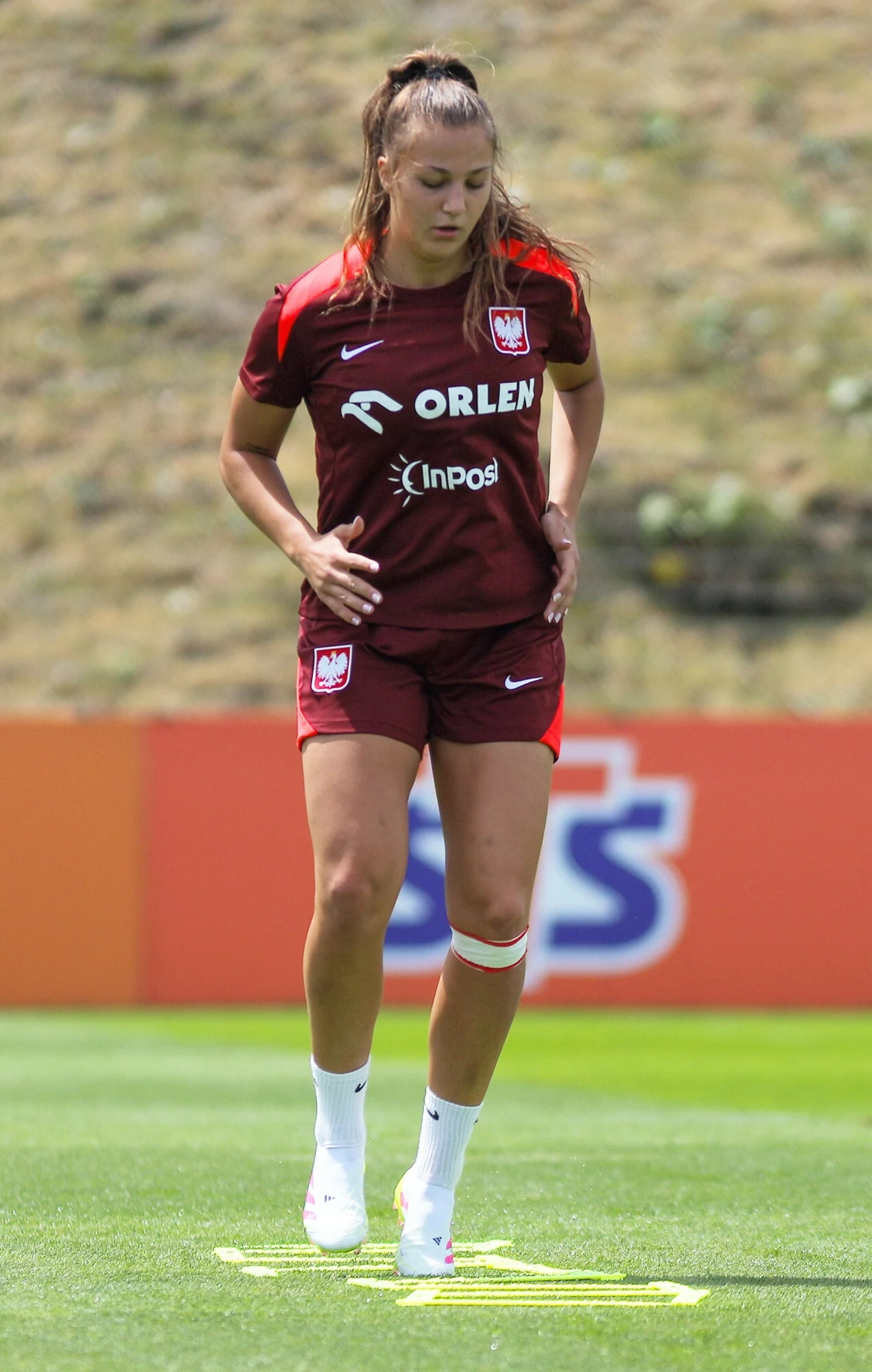 Emilia Szymczak Piłkarka w bordowym stroju reprezentacji Polski wykonuje ćwiczenie koordynacyjne na boisku treningowym, koncentrując się na ruchu nóg wokół oznaczeń na trawie.