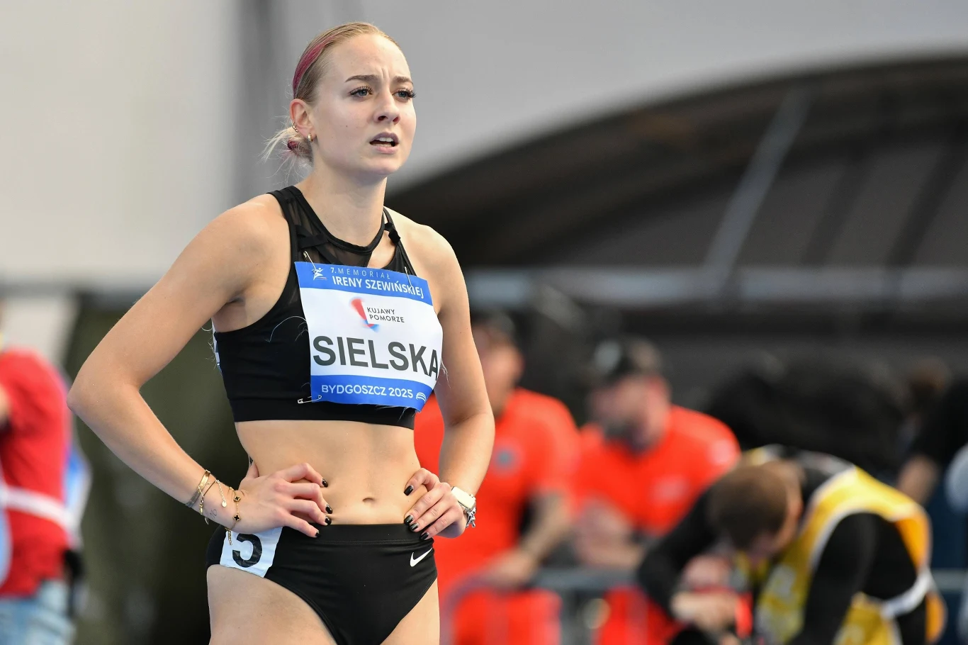 Alicja Sielska Lekkoatletka w sportowym stroju z numerem startowym na piersi, stojąca na bieżni z profesjonalnym wyrazem twarzy, w tle rozmyte sylwetki osób ubranych w pomarańczowe stroje organizacyjne.