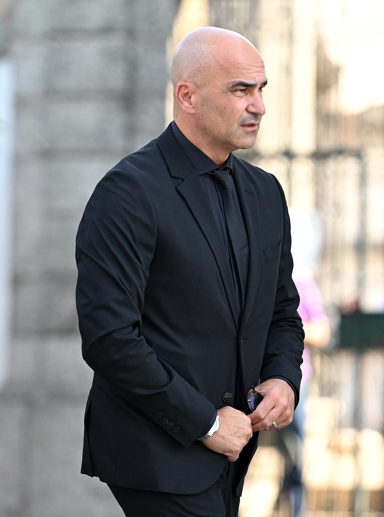 Roberto Martinez na pogrzebie Diogo Joty i Andre Silvy Mężczyzna w średnim wieku o łysiejącej głowie ubrany w czarny garnitur i czarną koszulę, stoi na zewnątrz w słoneczny dzień na tle rozmytej architektury.