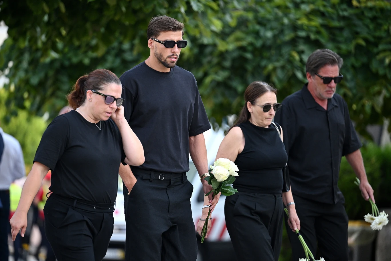 Czworo dorosłych ubranych na czarno idzie ulicą, trzymając białe kwiaty, z wyraźnym smutkiem na twarzach, co sugeruje udział w ceremonii żałobnej.