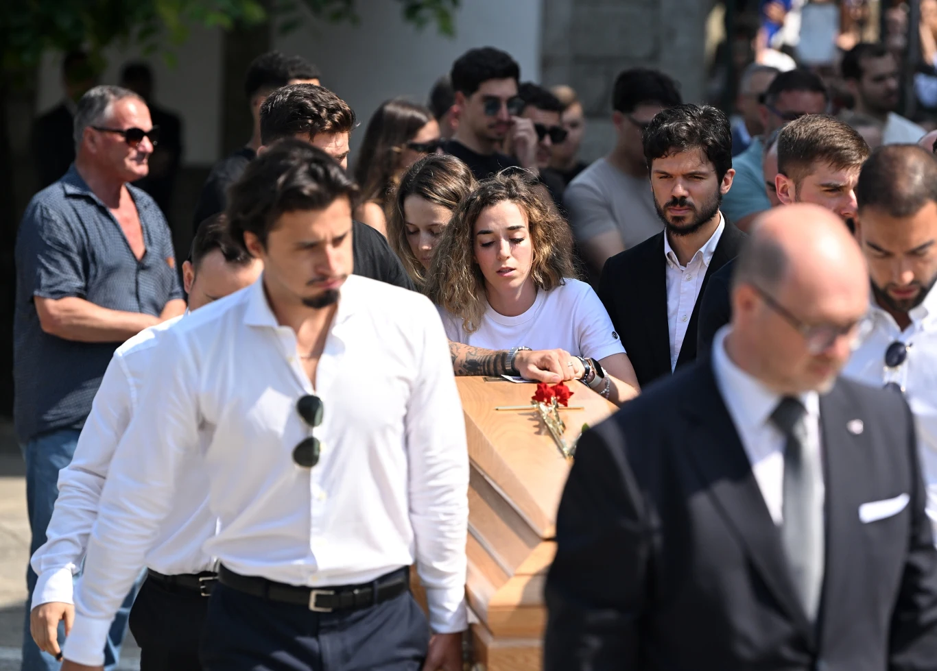 Grupa osób ubranych w ciemne ubrania idzie za drewnianą trumną, którą niesie kilku mężczyzn, jedna z kobiet opiera dłoń na trumnie, wokół panuje atmosfera powagi i smutku, na trumnie leży czerwona róża.