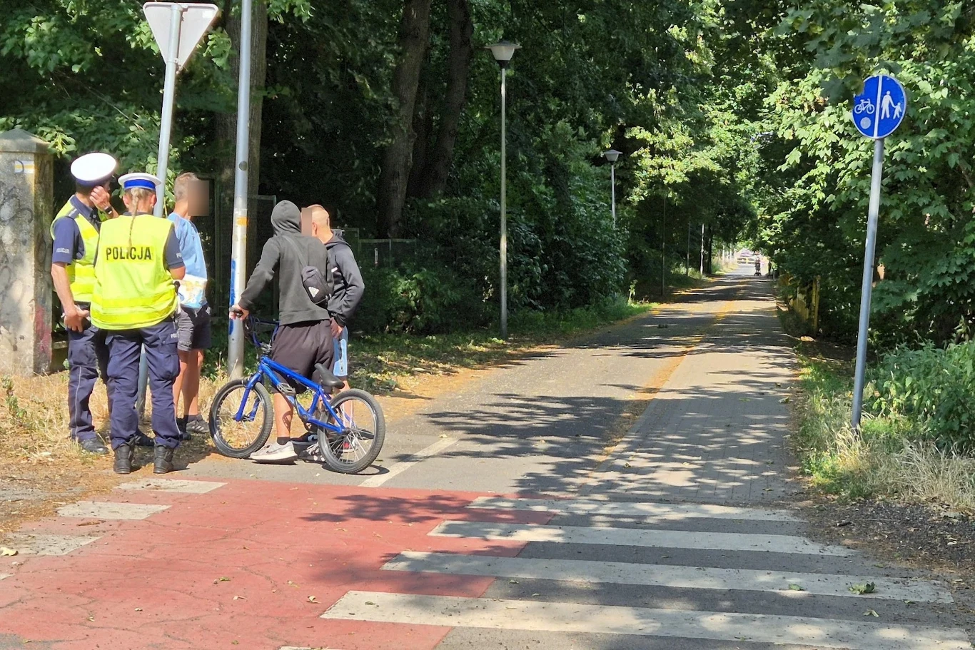 Policja uświadamiała rowerzystom obowiązujące ich przepisy ruchu Dwóch policjantów rozmawia z młodą osobą na rowerze, stojącą na przejściu dla pieszych i ścieżce rowerowej otoczonej zielenią; pogoda jest słoneczna, w tle prowadzi długa, zacieniona ścieżka.