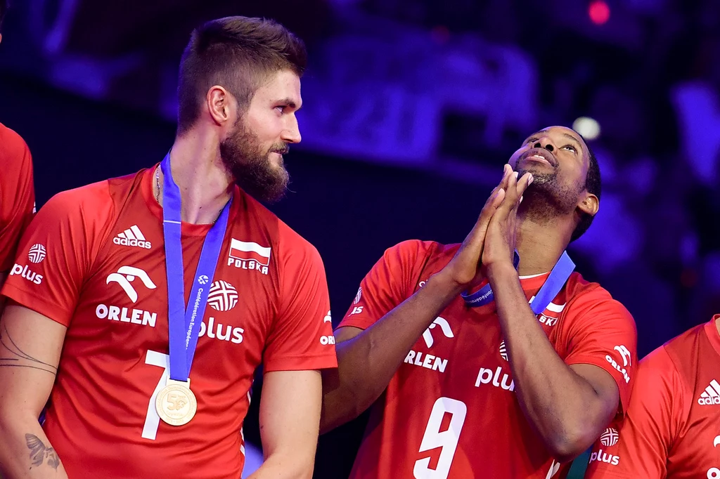 Karol Kłos i Wilfredo Leon Dwóch siatkarzy polskiej reprezentacji w czerwonych koszulkach z medalami na szyi, jeden z nich trzyma dłonie złożone w geście podziękowania lub modlitwy, drugi z uśmiechem spogląda na kolegę; w tle widownia i sportowe oświetlenie.