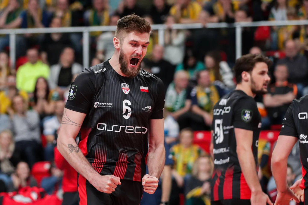 Karol Kłos Siatkarz w czarno-czerwonym stroju drużyny Asseco Resovia z okrzykiem radości i zaciśniętymi pięściami, wyrażający silne emocje na tle rozmytego tłumu kibiców.