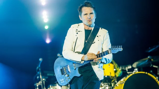 Muzyk w białej kurtce gra na niebieskiej gitarze elektrycznej na oświetlonej scenie koncertowej, w tle widoczna żółta perkusja i elementy wyposażenia sceny.