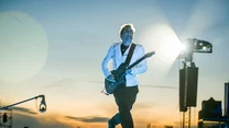 Mężczyzna w białej kurtce grający na gitarze elektrycznej na scenie podczas koncertu plenerowego o zachodzie słońca, w tle oświetlenie scena i zarysy publiczności.