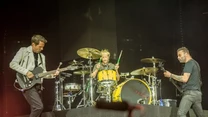 Trzech muzyków na scenie podczas koncertu, gitarzysta i basista po bokach, perkusista pośrodku za dużym żółtym zestawem perkusyjnym, kolorowe światła i scena podkreślają atmosferę występu na żywo.