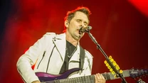 Muzyk w białej kurtce gra na elektrycznej gitarze i śpiewa do mikrofonu podczas koncertu, scena oświetlona intensywnym czerwonym światłem.
