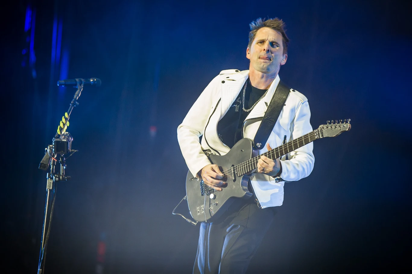 Muse na Open'er Festivalu 2025 Muzyk na scenie grający na elektrycznej gitarze, ubrany w białą kurtkę i ciemne spodnie, dynamiczne niebieskie oświetlenie oraz mikrofon po lewej stronie kadru.