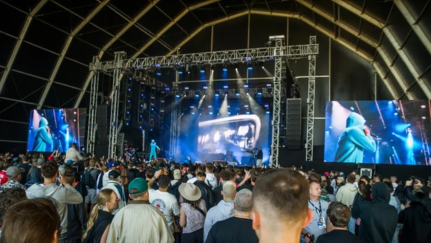 Tłum ludzi zgromadzony pod dużą sceną koncertową wewnątrz hali, scena oświetlona niebieskimi i białymi reflektorami, na bocznych ekranach widoczny jest występujący artysta, publiczność skupiona na występie.