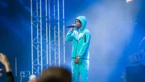 Młody mężczyzna w jasnoniebieskim dresie i kapturze wykonuje utwór na scenie podczas koncertu, trzymając mikrofon, wokół niego są światła sceniczne oraz widownia z podniesionymi rękami i telefonami.