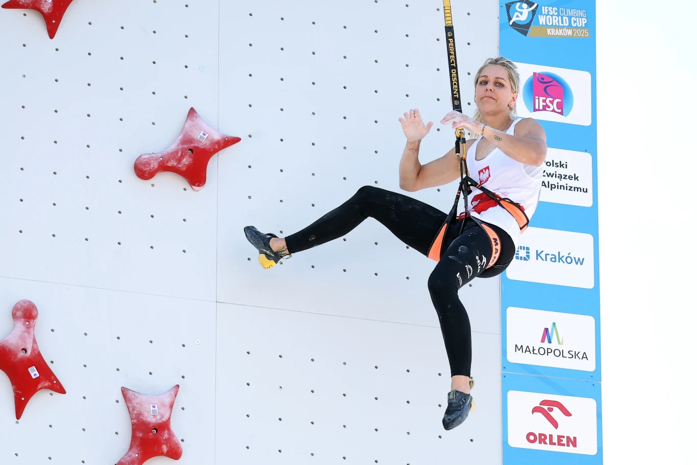 Aleksandra Mirosław Zawodniczka wspinaczki sportowej w uprzęży zahaczona o linę bezpieczeństwa podczas zawodów, tło stanowi ścianka wspinaczkowa oraz logotypy sponsorów i organizatorów imprezy.