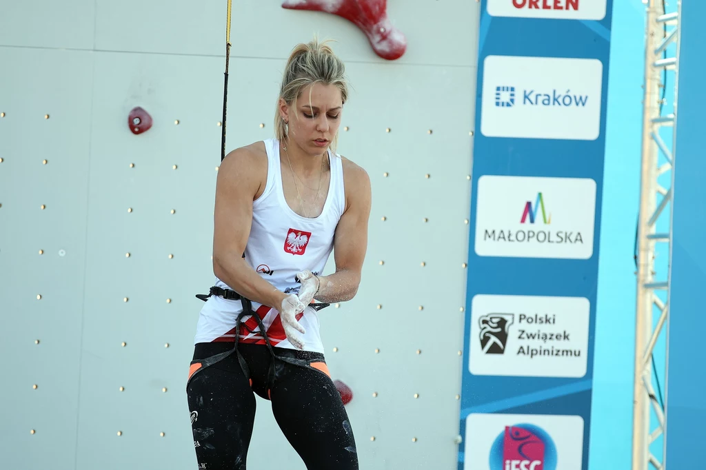 Aleksandra Mirosław Kobieta w sportowym stroju z emblematem Polski na koszulce, przygotowuje się do wspinaczki na ściance wspinaczkowej, posypując dłonie magnezją. W tle widoczne logotypy sponsorów i organizatorów zawodów.