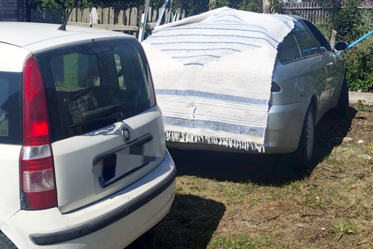 Takie maskowanie na niewiele się zdało Dwa zaparkowane samochody, jeden zasłonięty dywanem, drugi odsłonięty, ustawione na ziemistym podwórku przy drewnianym płocie.