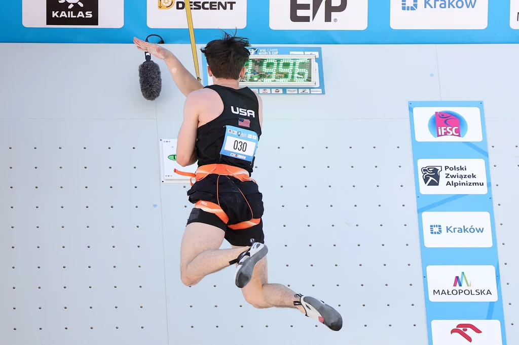 Samuel Watson Zawodnik reprezentujący USA w trakcie dynamicznej wspinaczki sportowej, wykonujący ruch w kierunku górnej krawędzi ścianki, widoczny numer startowy, elektroniczny zegar pomiaru czasu oraz logotypy sponsorów i miast partnerujących zawodom.
