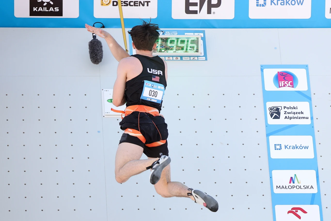 Samuel Watson Zawodnik reprezentujący USA w trakcie dynamicznej wspinaczki sportowej, wykonujący ruch w kierunku górnej krawędzi ścianki, widoczny numer startowy, elektroniczny zegar pomiaru czasu oraz logotypy sponsorów i miast partnerujących zawodom.
