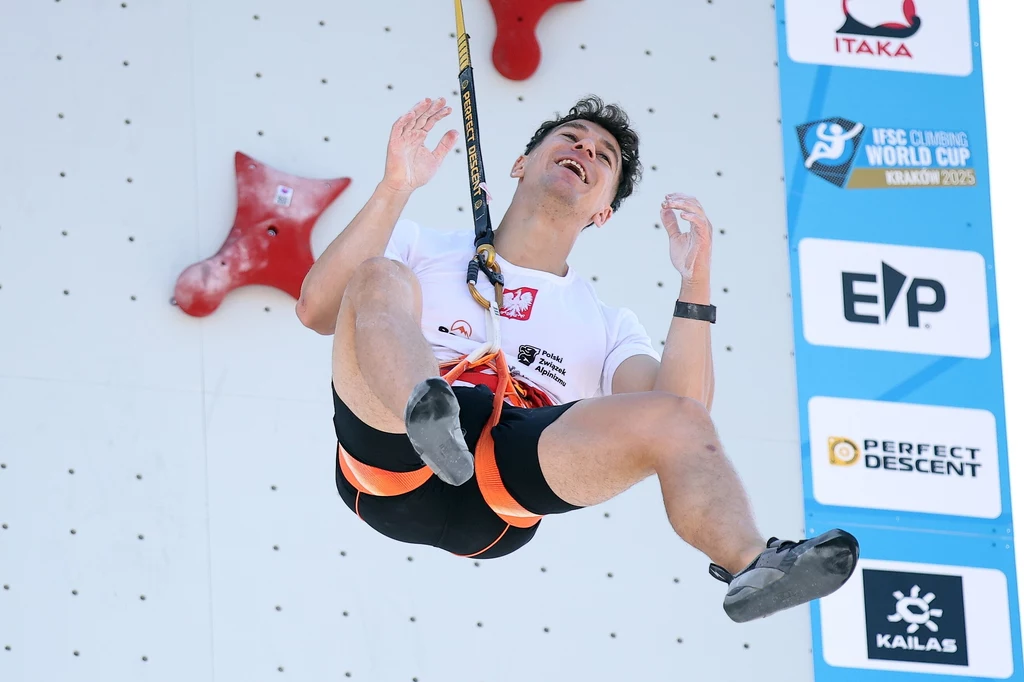 Marcin Dzieński Sportowiec w uprzęży wspinaczkowej unoszący się w powietrzu podczas zawodów wspinaczkowych na ścianie z widocznymi uchwytami oraz logotypami sponsorów i nazwą wydarzenia.