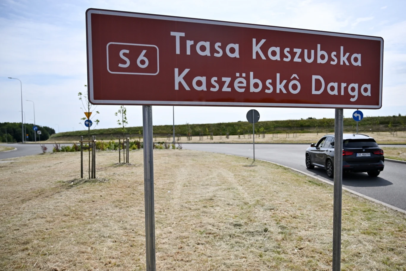 Dwujęzyczna tablica odsłonięta w sobotę na węźle Kamień - Południe Czerwony znak drogowy z napisem 'Trasa Kaszubska' w języku polskim i kaszubskim, oznaczający trasę S6 przy rondzie, po prawej stronie czarne auto wjeżdżające na rondo, w tle zieleń i niebo.
