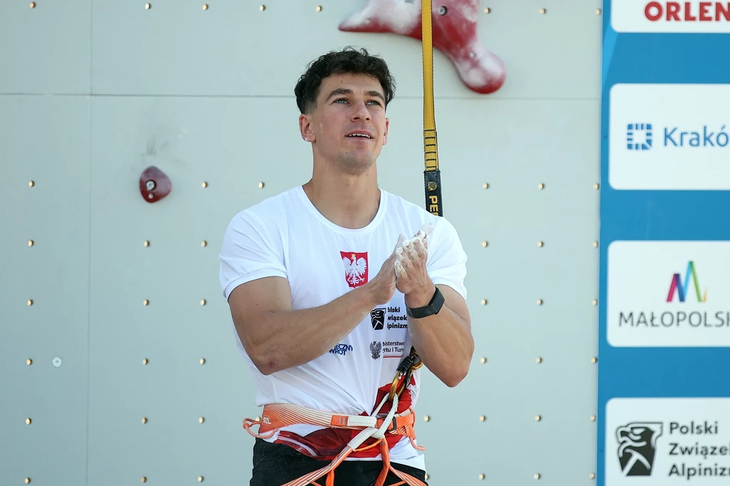 Sportowiec ubrany w białą koszulkę z godłem Polski i uprząż wspinaczkową stoi przy ścianie wspinaczkowej z charakterystycznymi chwytami i oznaczeniami sponsorów oraz organizatorów zawodów.