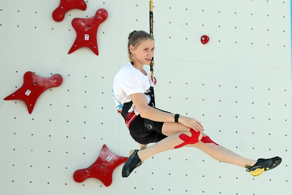 Aleksandra Kałucka Młoda kobieta w sportowym stroju wspinaczkowym, wisząca na uprzęży przy ściance wspinaczkowej, z widocznymi elementami chroniącymi kolano oraz charakterystycznymi czerwonymi chwytami na ścianie.