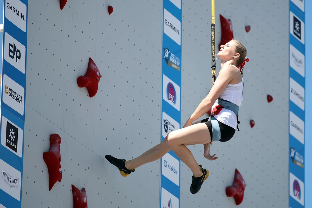 Natalia Kałucka Zawodniczka wspinaczki sportowej w uprzęży, zawieszona na linie przy ściance wspinaczkowej z czerwonymi chwytami, na tle jasnej ściany z widocznymi logotypami sponsorów.