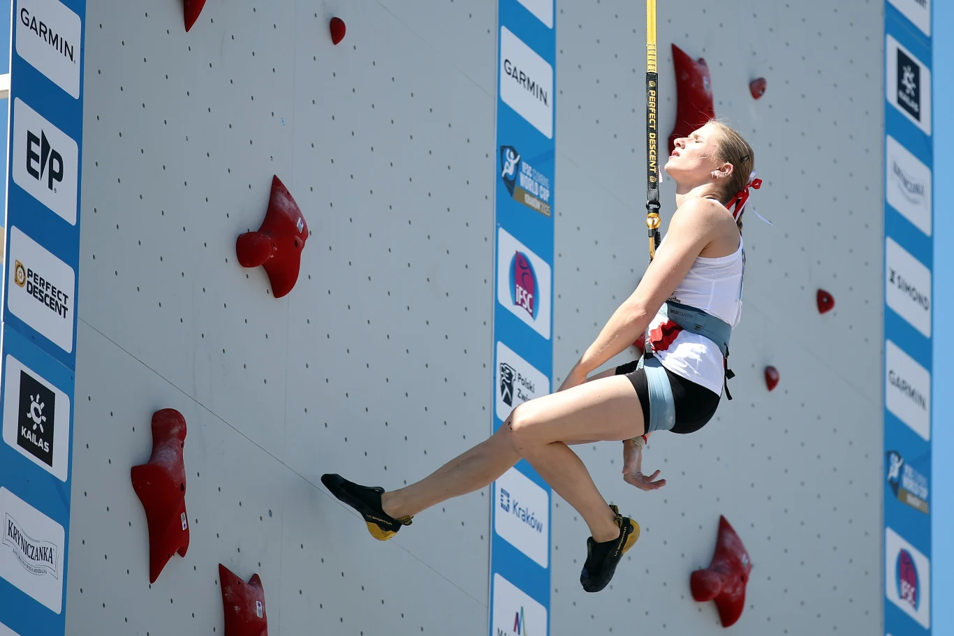 Natalia Kałucka Zawodniczka wspinaczki sportowej w uprzęży, zawieszona na linie przy ściance wspinaczkowej z czerwonymi chwytami, na tle jasnej ściany z widocznymi logotypami sponsorów.