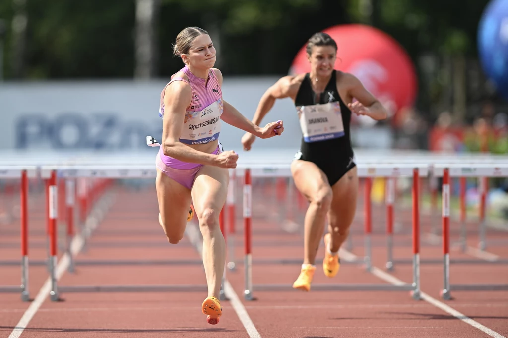 Pia Skrzyszowska w Poznaniu Dwie kobiety biorą udział w biegu przez płotki na stadionie lekkoatletycznym, skoncentrowane na przekroczeniu linii mety, w tle widoczna jest czerwona i niebieska konstrukcja oraz rozmazane elementy otoczenia.