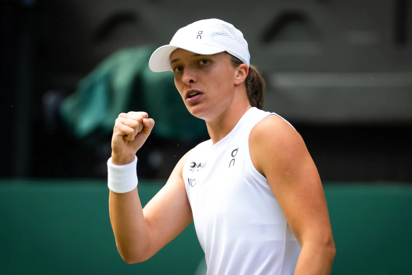Iga Świątek rywalizowała z Danielle Collins o awans do czwartej rundy Wimbledonu Tenisistka w białym stroju sportowym z czapką wykonuje gest zaciśniętej pięści, wyrażający determinację i zwycięstwo. W tle rozmazane elementy kortu tenisowego.