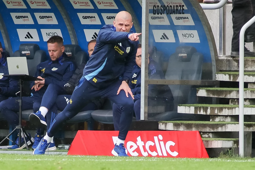 Dawid Szwarga Trener drużyny piłkarskiej energicznie gestykuluje i wskazuje kierunek podczas meczu, siedząc na ławce rezerwowych wraz z innymi członkami sztabu obok stadionowej murawy.